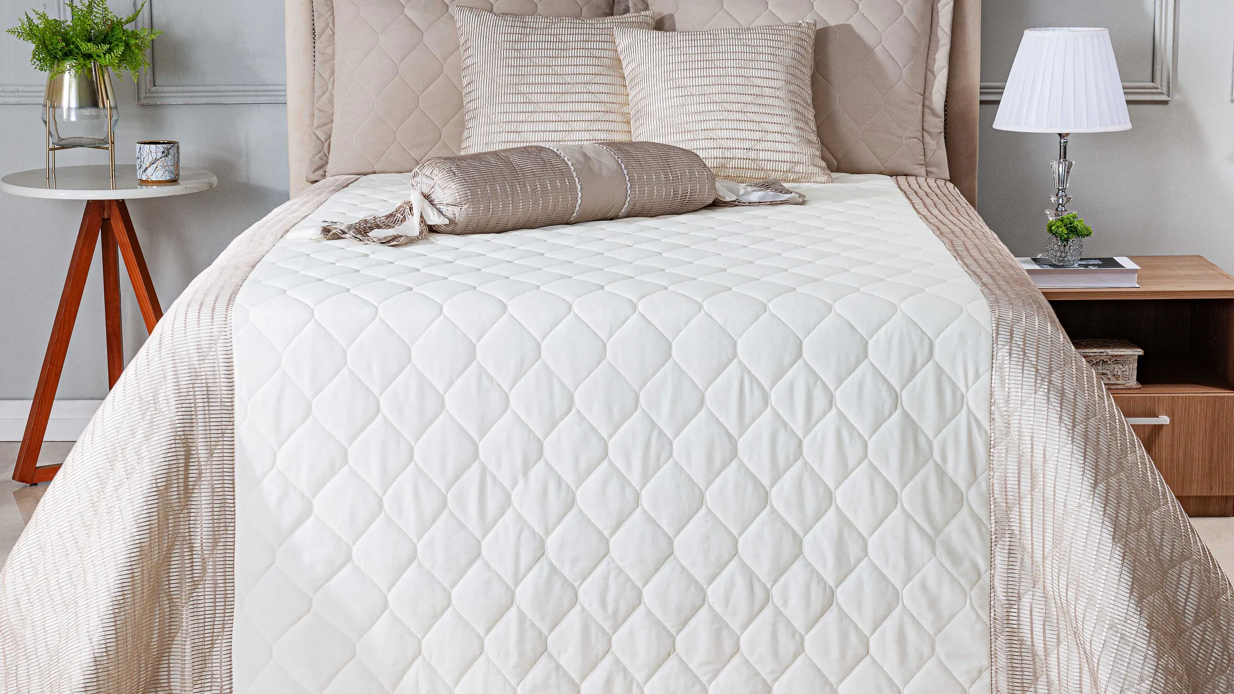 Cama de casal montada com colcha matelassê branca, almofadas e rolinho bordados, num quarto claro